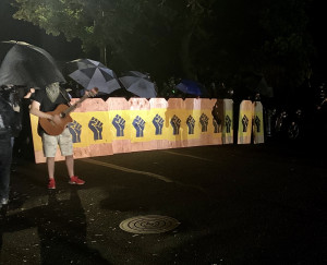 A row of wooden shields each decorated with black fists on a yellow background. The shields form a shield wall with a row of umbrellas above them. A person playing the guitar stands in front of the shield wall to the side. 