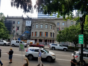 People gathered at a protest across the street from Portland city hall with Pride flags in the windows.