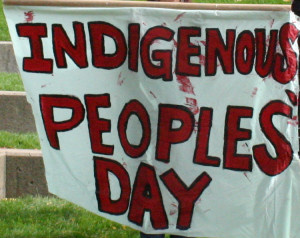 Hand-painted banner, waving slightly in wind, red letters on white background,  saying "Indigenous People's Day"