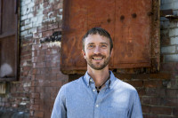 Jason Rasor wearing a blue shirt and jewelry smiles while standing in front of a brick wall with rusted metal sections.