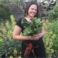Marisha Auerbach, wearing a black dress and pruning tool belt, smiles while holding a bouquet of fresh cut greens